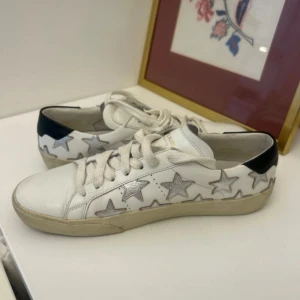 Vita sneakers med stjärnor från Saint Laurent - Snygga vita sneakers från Saint Laurent med silverstjärnor och svarta detaljer på hälen. Nypris cirka 6000. Bra skick inte använt så många gånger:)