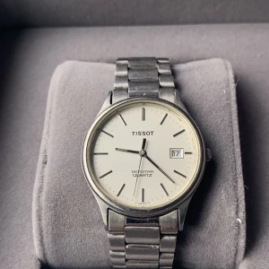 Tissot Seastar Quartz klocka - Elegant Tissot Seastar Quartz klocka med en stilren vit urtavla och datumvisning. Klockan har en klassisk design med ett silverfärgat armband i rostfritt stål. Perfekt för den som söker en tidlös accessoar.