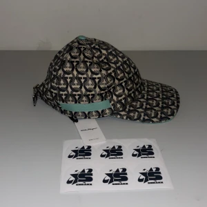 Mönstrad keps från Salvatore Ferragamo - Snygg keps med ett unikt mönster i svart och beige från Salvatore Ferragamo. Kepsen har en justerbar rem baktill och en stilren design med en touch av turkos på skärmen och remmen. Perfekt för att ge din outfit en exklusiv känsla.