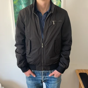 Svart bomberjacka från Riley - Snygg svart bomberjacka från Riley med dragkedja och ribbade muddar. Jackan har en bröstficka med dragkedja och två sidofickor. Perfekt för en stilren look. NyPris ligger på cirka 1999kr😊