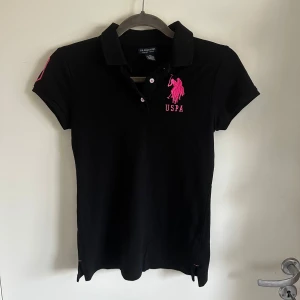 Svart pikétröja från U.S. Polo Assn. - Polo ralph lauren liknande pikétröja.  svart pikétröja från U.S. Polo Assn. med rosa broderad logga på bröstet och siffran 3 på ärmen. Tröjan har korta ärmar och knappar framtill. Pris kan diskuteras💞 