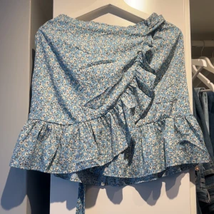 Blommig blå omlottkjol - Söt blå omlottkjol med blommigt mönster och volangdetaljer. Kjolen har en knytning i midjan. 