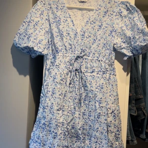 Blommig klänning med knytband - Säljer en söt blommig klänning i vitt och blått med korta puffärmar och v-ringning. Klänningen har ett knytband i midjan för en fin passform. 