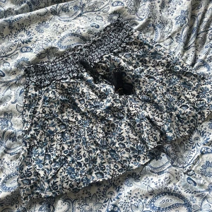Blommig kortkjol med volang - Charmig kortkjol med blommigt mönster i blått och vitt. Kjolen har en elastisk midja med volangdetaljer och ett knytband framtill. Perfekt för en somrig look!