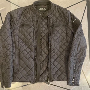 Svart quiltad jacka från Replay - Snygg svart quiltad jacka från Replay med dragkedja och knappar. Jackan har långa ärmar och flera praktiska fickor med dragkedjor. Perfekt för en stilren look. Smal i storleken.