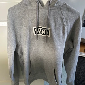 Grå hoodie från Vans - Säljer en grå hoodie från Vans med klassisk logga på bröstet. Tröjan har en känguruficka och snörning i huvan. På ärmen finns ett svartvitt rutmönster som ger en cool touch. Perfekt för en avslappnad stil.