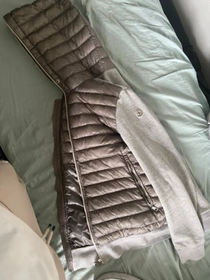Grå dunjacka med huva från Moncler - Snygg grå dunjacka från Moncler med quiltad front och huva. Ärmarna är i mjukare material och jackan har dragkedja framtill samt fickor med dragkedja. Perfekt för kalla vinterdagar och har en modern, sportig look.