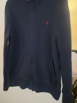 Mörkblå ziptröja från Polo Ralph Lauren - Mörkblå stickad ziptröja från Polo Ralph Lauren med röd broderad logga på bröstet. Tröjan har två framfickor, ribbade muddar och hög krage. Perfekt för lager på lager och en clean look.