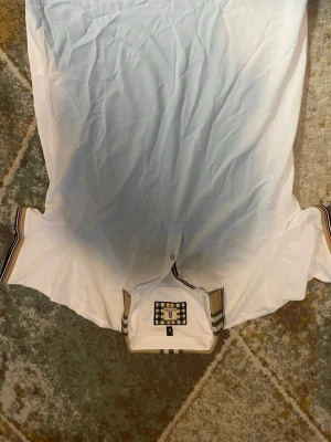 Vit skjorta med rutiga detaljer - Snygg vit skjorta med klassiska rutiga detaljer på kragen och insidan av knappslån. Skjortan har korta ärmar och en stilren design som passar till många olika outfits. Materialet ser ut att vara bomull och skjortan har en modern passform.