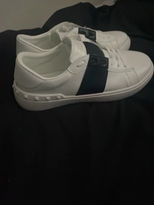 Valentino - Säljer ett par vita sneakers med bred svart rem över sidan och coola nitar på sulan. Skorna har klassisk låg profil och vita snören. Perfekta för dig som vill ha en clean men ändå edgy stil.