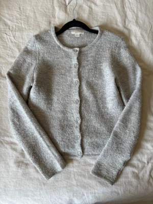 Ljusgrå fluffig kofta med knappar - Mysig ljusgrå kofta med rund halsringning och knäppning framtill. Koftan har en fluffig, mjuk struktur och långa ärmar. Enkel och stilren design som passar till många olika looks.