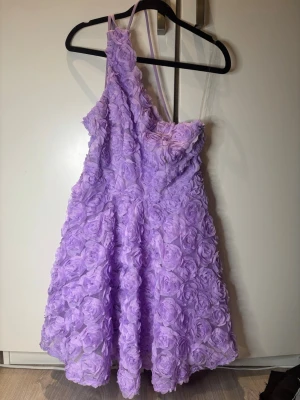 Ljuslila one shoulder klänning - Superfin ljuslila klänning från one shoulder-design och 3D-blommor över hela tyget. Klänningen är kort och har en voluminös kjol som ger en romantisk look. Perfekt för dig som vill sticka ut med en unik och söt stil. Använd 1 gång