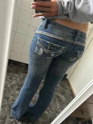 Blå bootcut jeans med detaljer - Säljer ett par blå bootcut jeans med slitningar och dekorativa sömmar samt fickdetaljer bak. Jeansen har låg midja och coola tvättade effekter som ger en trendig look. Perfekta för dig som gillar en avslappnad men ändå snygg stil.