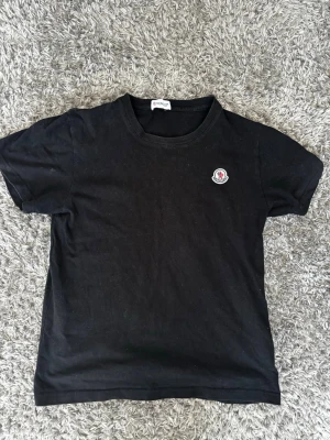 Svart Moncler t-shirt med logga - Säljer en svart t-shirt från Moncler med klassisk rund hals och korta ärmar. På bröstet sitter Monclers ikoniska logga i vitt, blått och rött. T-shirten är i mjuk bomull och har en clean, stilren look som passar till allt.                        Storlek S