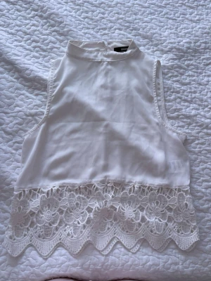 Vit ärmlös topp med spetsdetalj - Säljer en vit ärmlös topp med hög krage och vacker spetskant nedtill. Toppen har en rak passform och stängs med en liten knapp i nacken. Spetsen har ett blommigt mönster och ger toppen en romantisk touch. Perfekt till sommaren! Denne genomskinlig oxå