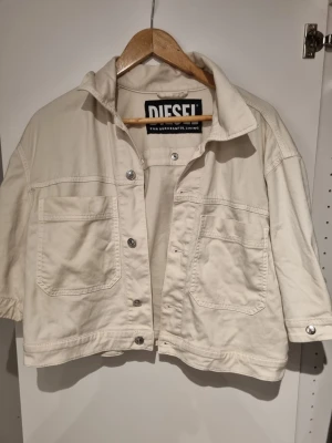 Diesel D-Reef-Ne Jacket  - Säljer en vit jeansjacka från Diesel med coola fransdetaljer och silverfärgade knappar. Jackan har två stora bröstfickor, kortare passform och unika fransar på ryggen. Perfekt för dig som vill sticka ut med en trendig och edgy look.