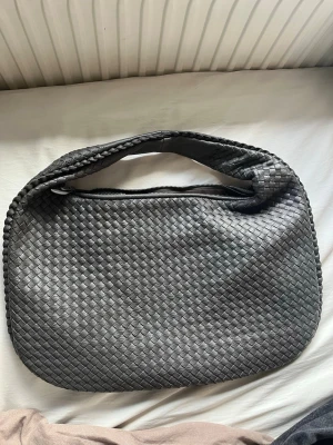 Svart flätad axelväska i skinnimitation - Snygg mörkgrå väska med flätad design och rundad form. Väskan har dragkedja upptill och ett rymligt innerfack med extra fickor, perfekt för att hålla ordning på dina grejer. Materialet är skinnimitation och axelremmen är bred och bekväm.