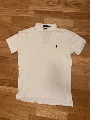 Vit pikétröja från Polo Ralph Lauren - Klassisk vit pikétröja från Polo Ralph Lauren med blå logga broderad på bröstet. Tröjan har korta ärmar, krage och knappar framtill. Modellen är slim fit och tillverkad i bomull för en skön känsla.