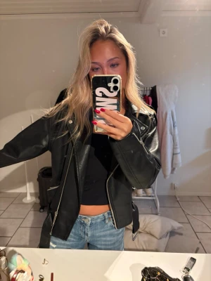 Svart  skinnjacka - Säljer en svart skinnjacka i klassisk bikerstil med sned dragkedja, breda slag och silverfärgade detaljer. Jackan har fickor med dragkedja och justerbara spännen vid ärmsluten. Även ett skärp. 