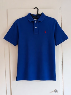 Ralph Lauren piké - Polo Ralph Lauren pikétröja i en snygg klarblå färg med det klassiska röda broderade Ralph Lauren-märket på bröstet. 💙❤️ Storlek L (14–16), vilket är en juniorstorlek. Tillverkad i 100% bomull. Klassisk modell med krage, två knappar och kort ärm. Fint skick utan defekter! Perfekt till både vardags och en mer dressad outfit. 📏 Storlek: L (14–16) 🧵 Material: 100% bomull 🏷️ Polo Ralph Lauren