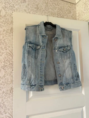 Ljusblå ärmlös jeansjacka Unique Denim - Snygg ljusblå ärmlös jeansjacka från Unique Denim i storlek 38. Klassisk modell med krage, bröstfickor med lock och knappar framtill. Perfekt att slänga över en hoodie eller t-shirt för en cool och avslappnad look.