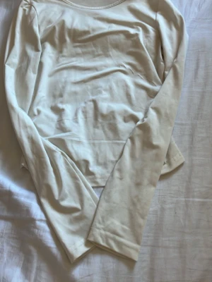 Ljusvit långärmad topp - En enkel och stilren ljusvit långärmad topp med rund halsringning. Toppen har en lätt rynkning i sidan som ger en snygg detalj. Tillverkad i mjukt material som känns skönt mot huden. Perfekt att matcha med jeans eller kjol.