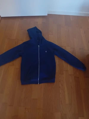 Mörkblå hoodie med dragkedja Polo - Snygg mörkblå hoodie från Polo Ralph Lauren med dragkedja framtill och klassisk vit logga på bröstet. Hoodien har huva och långa ärmar, perfekt för en chill och stilren look. Tillverkad i mjukt material som känns skönt mot huden.