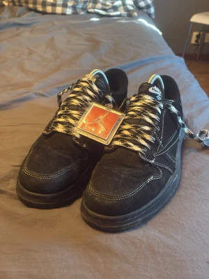 Nike Air Force 1 x Travis Scott svart - Säljer ett par Nike Air Force 1 x Travis Scott sneakers i svart mocka med vita kontrastsömmar och unika snören i svartvitt mönster. Skorna har en avtagbar silverfärgad detalj på snörningen och Travis Scott-detaljer på innersulan. Perfekta för dig som gillar streetwear.