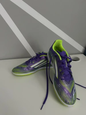 Lila och limegröna Adidas  inomhusskor  - Säljer ett par snygga Adidas fotbollsskor i lila och limegrönt med vita detaljer. Skorna har låg profil, snörning och en slitstark syntetisk ovandel. Perfekta för dig som vill sticka ut på planen med både stil och funktion.