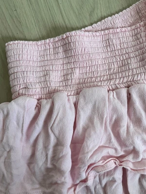 Ljusrosa volangkjol med smock - Supersöt ljusrosa kortkjol med bred smock i midjan och dubbla volanger. Kjolen är gjord i mjukt bomullstyg och har en luftig känsla som passar perfekt till varma dagar. Enkel att matcha med både toppar och sneakers för en chill look.