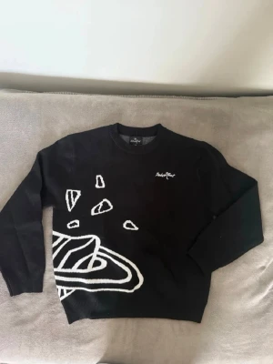 Broken planet - Cool svart stickad tröja med vita grafiska rymdmotiv som planeter, stjärnor och asteroider både fram och bak. Rund halsringning och ribbade muddar. Perfekt för dig som gillar unika och trendiga plagg med en twist.
