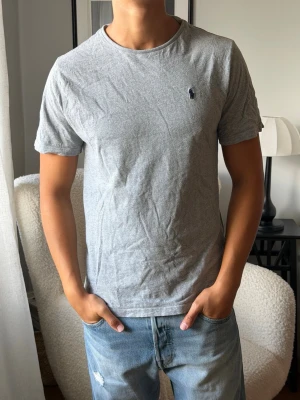 Grå t-shirt med liten logga - En enkel grå t-shirt i bomull med rund hals och korta ärmar. På bröstet finns en liten broderad logga i mörkblått. Passformen är normal och tyget känns mjukt och bekvämt, perfekt till jeans eller shorts. Är i L barn men sitter som en S herr