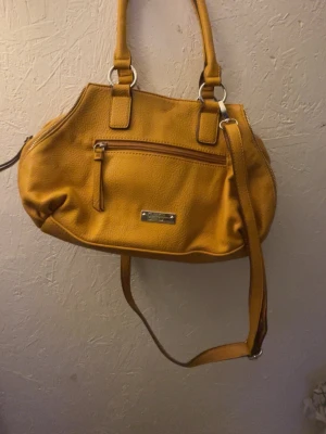 Gul handväska från Jessica Simpson - Snygg gul handväska från Jessica Simpson i mjukt konstläder. Väskan har både handtag och justerbar axelrem, samt en praktisk ytterficka med dragkedja och silverfärgade metalldetaljer. Perfekt för dig som vill ha en färgklick till din outfit.
