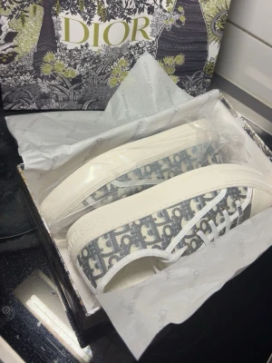 Gråvita Dior sneakers med monogram - Snygga låga sneakers från Dior i grått och vitt med klassiskt Dior-monogram över hela skon. Skorna har vita snören, robust vit sula och detaljer med Christian Dior-logga på sidan och hälen. Perfekta för dig som vill ha en exklusiv och trendig look.