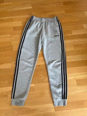 Grå Adidas mjukisbyxor med svarta ränder - Snygga grå mjukisbyxor från Adidas med klassiska svarta ränder längs benen och elastisk midja. Byxorna har ribbade muddar vid bensluten och är perfekta för chill eller träning. Adidas-logga på vänster lår. Mjukt och bekvämt material.