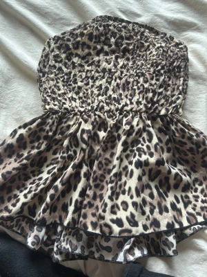 Leopardmönstrad bandeau topp från shein - Säljer en leopardmönstrad bandeau topp från shein i storlek XXS. Toppen är axelbandslös med smockad överdel och volangig nederkant. Perfekt för dig som vill sticka ut med ett djurmönster och gillar en croppad look.
