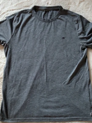 Mörkgrå t-shirt från Calvin Klein - En stilren mörkgrå t-shirt från Calvin Klein med diskret 'ck'-logga broderad på bröstet. T-shirten har rund halsringning och korta ärmar. Tillverkad i mjukt och tunt material som känns skönt mot huden. Men är som storlek S