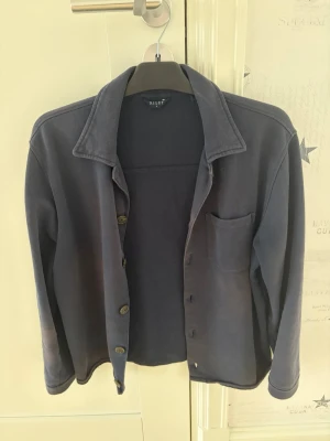 Riley overshirt  - Mörkblå/grå Riley-jacka – strl S  Snygg och stilren jacka från Riley i mörkblå/grå färg. Klassisk modell med skjortkrage, knappstängning framtill och praktisk bröstficka. Fina sömdetaljer på rygg och axlar samt en snygg, något figurnära passform.  Perfekt som vår-/höstjacka och passar lika bra till vardags som till en mer uppklädd outfit.  📏 Storlek: S 🎨 Färg: Mörkblå/grå ✨ Skick: Mycket fint skick  Pris: 349 kr  Skickas snabbt och packas väl 📦