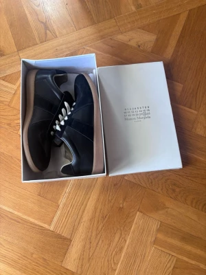 Maison margiela gats  - Snygga Maison Margiela gats svarta sneakers  med ovandel i mocka och läder. Klassisk gummisula och neutrala snören. Perfekta för dig som gillar märkeskläder, priset är svinbra pågrund av att de är köpta på Plick helt nya för 4000 och är lite osäker på äkthet och kan tyvärr inte garantera de. Det är testade och använd en kväll  men sen så köpte morsan ett par andra  som jag själv gillade mer så använde ej dessa mer, box ingår, skriv vid minsta fundering och fraktar vid samma dag  1599 superpris.
