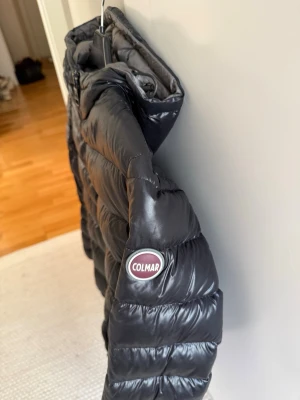 Svart dunjacka från Colmar - Snygg svart dunjacka från Colmar med glansig finish och huva. Jackan har quiltad design, dragkedja framtill och Colmar-logga på ärmen. Perfekt för kalla vinterdagar och riktigt skön att bära. Den har ett litet hål på ärmen som jag har satt en svart liten lagningslapp över, inget som syns om man inte vet om det. Jag har även tyvärr råkat få halloween blod på insidan av jackan på lapparna. 