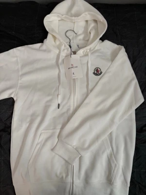 Vit hoodie med dragkedja från Moncler - Säljer en fräsch vit hoodie från Moncler med dragkedja framtill och klassisk Moncler-logga på bröstet. Hoodien har huva med dragsko och praktiska fickor på magen. Tillverkad i mjukt material som känns skönt mot huden. Perfekt för en clean och stilren look.