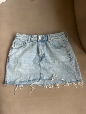 Ljusblå jeanskjol med fransig kant - Säljer en ljusblå jeanskjol i kort modell med rå, fransig kant nertill. Kjolen har klassiska fem fickor och bälteshällor. Perfekt för dig som gillar en avslappnad och trendig stil.
