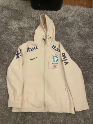 Vit Brasil Nike vindjacka med huva - Snygg vit vindjacka med huva och dragkedja framtill. Jackan har Brasilien-logga, Nike-logga och tryck på ärmarna i blått. Perfekt för dig som gillar sportig stil och vill sticka ut med landslagskänsla.
