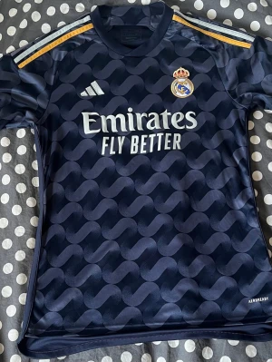 Mörkblå Real Madrid matchtröja - Snygg mörkblå Real Madrid matchtröja från Adidas med gulddetaljer på axlarna och klubbmärke på bröstet. Tröjan har ett vågigt mönster, korta ärmar och Aeroready-material som andas. Perfekt för fotbollsträning eller att bära till vardags.