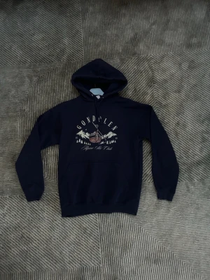 Mörkblå hoodie från blnc studio  - Säljer en mörkblå hoodie med huva från blnc studio och magficka. På bröstet finns ett tryck med texten 'Gondolen' och motiv av berg och en linbana. Hoodien har ribbade muddar vid ärmar och nederkant. Perfekt för dig som gillar avslappnad stil.