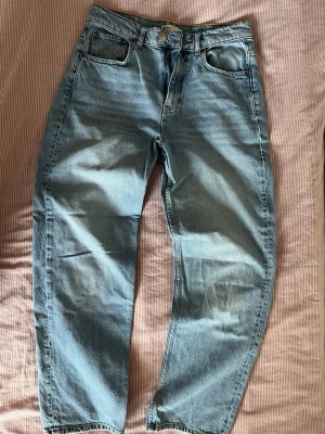 Ljusblå jeans - Säljer ett par ljusblå jeans med rak passform och klassisk femficksdesign. Jeansen har medel hög midja och är tillverkade i denim med snygga slitningar och tvättade detaljer. Perfekta för en avslappnad och trendig look.