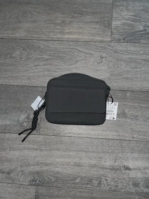 Svart/Grå axelväska från Pull&Bear - Snygg svart axelväska från Pull&Bear med minimalistisk design. Väskan har en framficka, dragkedja upptill och justerbar axelrem med flätad detalj. Tillverkad i ett slätt, matt material som ger en modern look. Perfekt för att bära det viktigaste.
