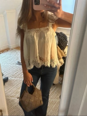Ljus spetsblus med volangdetaljer - Romantisk ljus blus med spetsdetaljer och volangkant. Blusen har breda axelband och en lös, luftig passform. Perfekt för dig som gillar en feminin och bohemisk stil. Materialet känns lätt och svalt, och spetsen ger en extra söt touch.