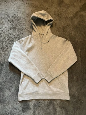 Ljusgrå hoodie med huva - En oanvänd stilren ljusgrå hoodie med huva och dragsko. Hoodien har en klassisk känguruficka framtill och ribbade muddar vid ärmslut och nederkant. Passformen av hoodien är relativt slim och tillverkad i mjukt sweatshirtmaterial. Pris går att diskutera!