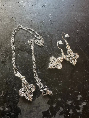 Silverfärgat halsband med lilja-hänge - Säljer ett silverfärgat halsband med ett detaljerat liljaformat hänge i klassisk fleur-de-lis-design. Kedjan är tunn och smidig, och hänget har fina ornamenterade detaljer. Perfekt för dig som gillar stilrena och tidlösa accessoarer.
